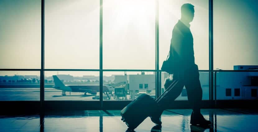 Fotografia przedstawia mężczyznę idącego z czarną walizką przez terminal na lotnisku. Mężczyzna ubraný jest w garnitur i rozgląda się spiesznie na boki, jakby zmierzając na samolot. Ruch postaci oraz otoczenie kojarzące się z lotniskiem nawiązują do tematyki podróży służbowych za granicę. Zdjęcie może ilustrować artykuły lub poradniki na ten temat.
