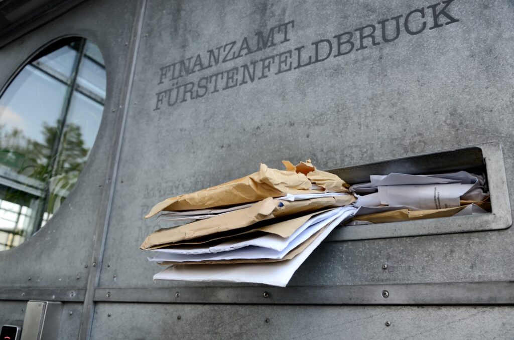 Fotografia przedstawia otwartą skrzynkę pocztową w kolorze żółtym z czerwonym daszkiem. Na skrzynce umieszczono duży, biały napis "Finanzamt". W otworze do wrzucania listów widać koperty i złożone kartki - są to dokumenty adresowane do urzędu skarbowego w Niemczech.