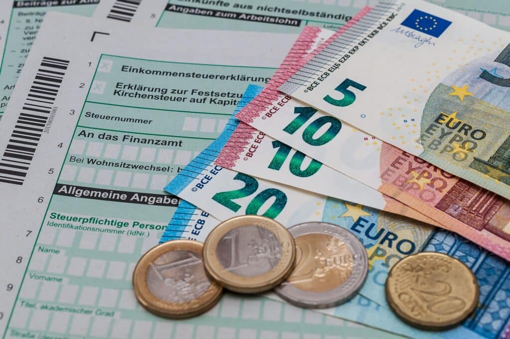 Fotografia przedstawia banknoty euro o nominałach 10 i 20 euro leżące na tle dokumentów skarbowych. Widać kilka banknotów ułożonych jeden na drugim. Pod nimi znajdują się formularze i wydruki w języku niemieckim dotyczące rozliczeń